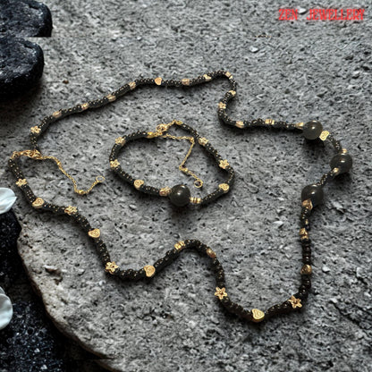 The jewellery set is photographed against a natural stone-inspired surface that complements the organic beauty of the black agate gemstones. The neutral grey tones provide excellent contrast, allowing the deep black beads and warm gold details to stand out clearly. This styling demonstrates how the jewellery combines bohemian influences with refined elegance suitable for both everyday outfits and special occasion wear.
