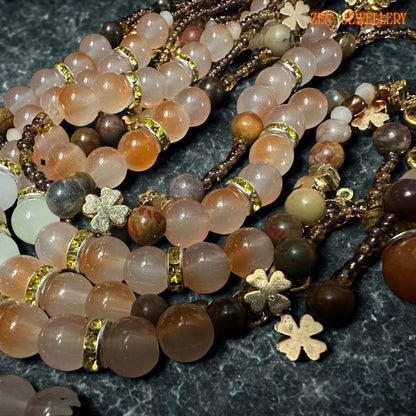 Romantic handmade gemstone necklace arranged on textured dark stone background featuring layered strands of peach, beige, caramel, and warm brown aventurine beads accented with sparkling champagne crystal beads. Gold floral charms and a celestial sun pendant add mystical artistic detail while the bracelet complements the necklace with matching colours and an adjustable clasp design.