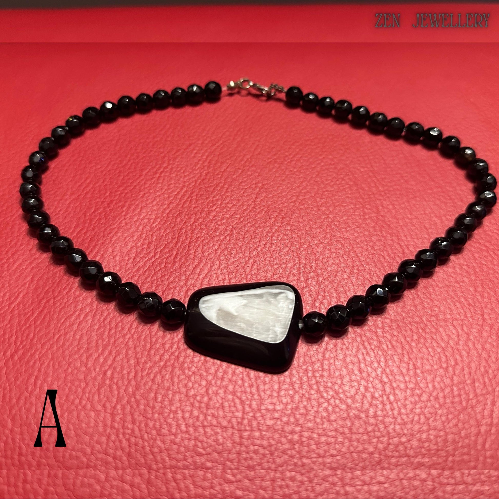 Handmade black crystal jewellery set photographed on a textured deep red surface to highlight the contrast between glossy black faceted beads and the iridescent white geometric pendant centre. The necklace strand curves naturally across the image, showing the fluid flexible construction and elegant arrangement of the polished beads. The jewellery design appears bold yet refined, ideal for women who love artistic statement accessories and contempo