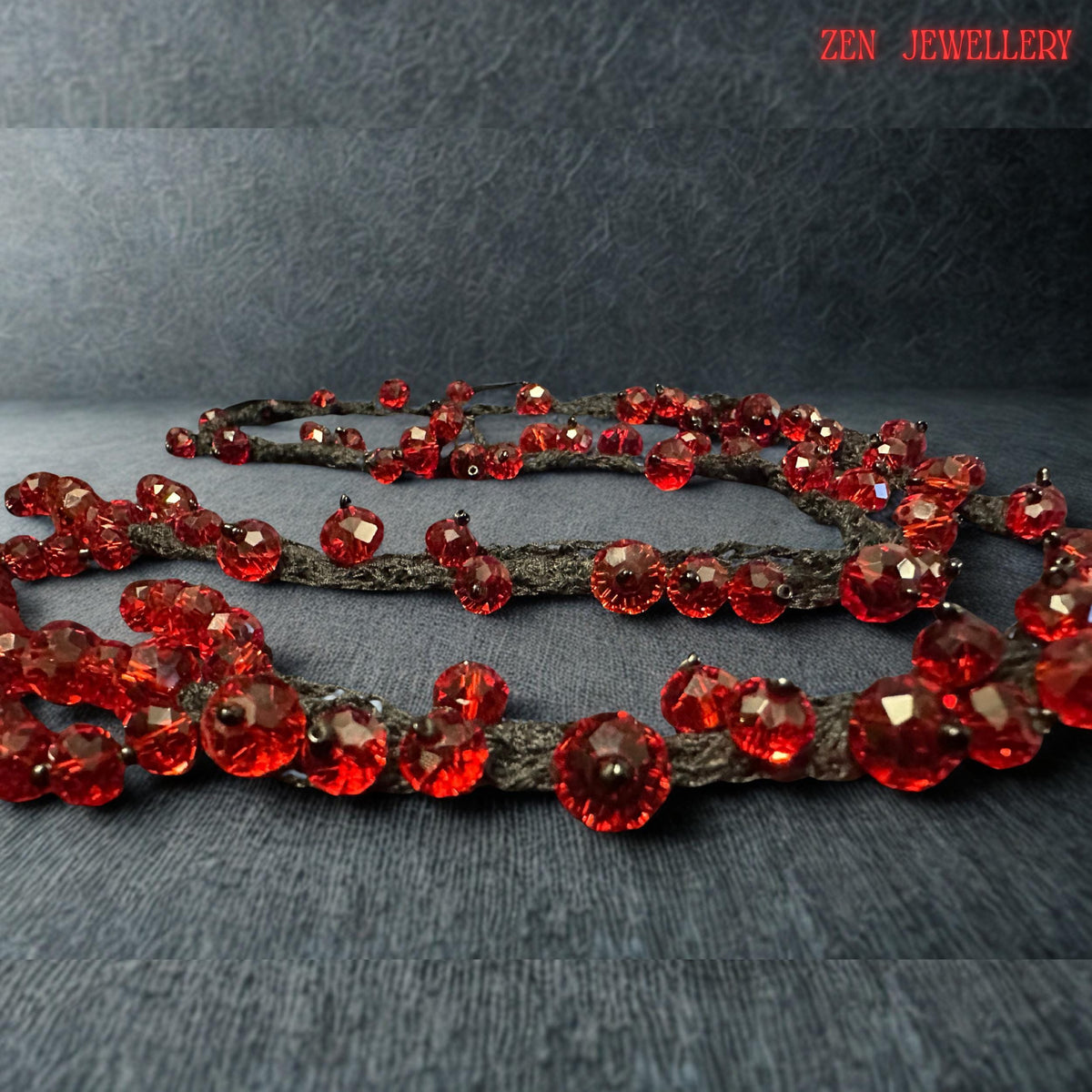 Romantic red crystal jewellery design displayed on a textured stone-style surface to emphasise the luxurious shine of the faceted scarlet beads. The handcrafted necklace features flowing black crochet strands decorated with vibrant ruby crystals in multiple sizes for added depth and movement. The bold artistic structure creates a statement appearance while remaining lightweight and flexible for comfortable wear. The dark base enhances the brillia