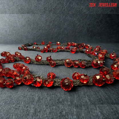 Romantic red crystal jewellery design displayed on a textured stone-style surface to emphasise the luxurious shine of the faceted scarlet beads. The handcrafted necklace features flowing black crochet strands decorated with vibrant ruby crystals in multiple sizes for added depth and movement. The bold artistic structure creates a statement appearance while remaining lightweight and flexible for comfortable wear. The dark base enhances the brillia