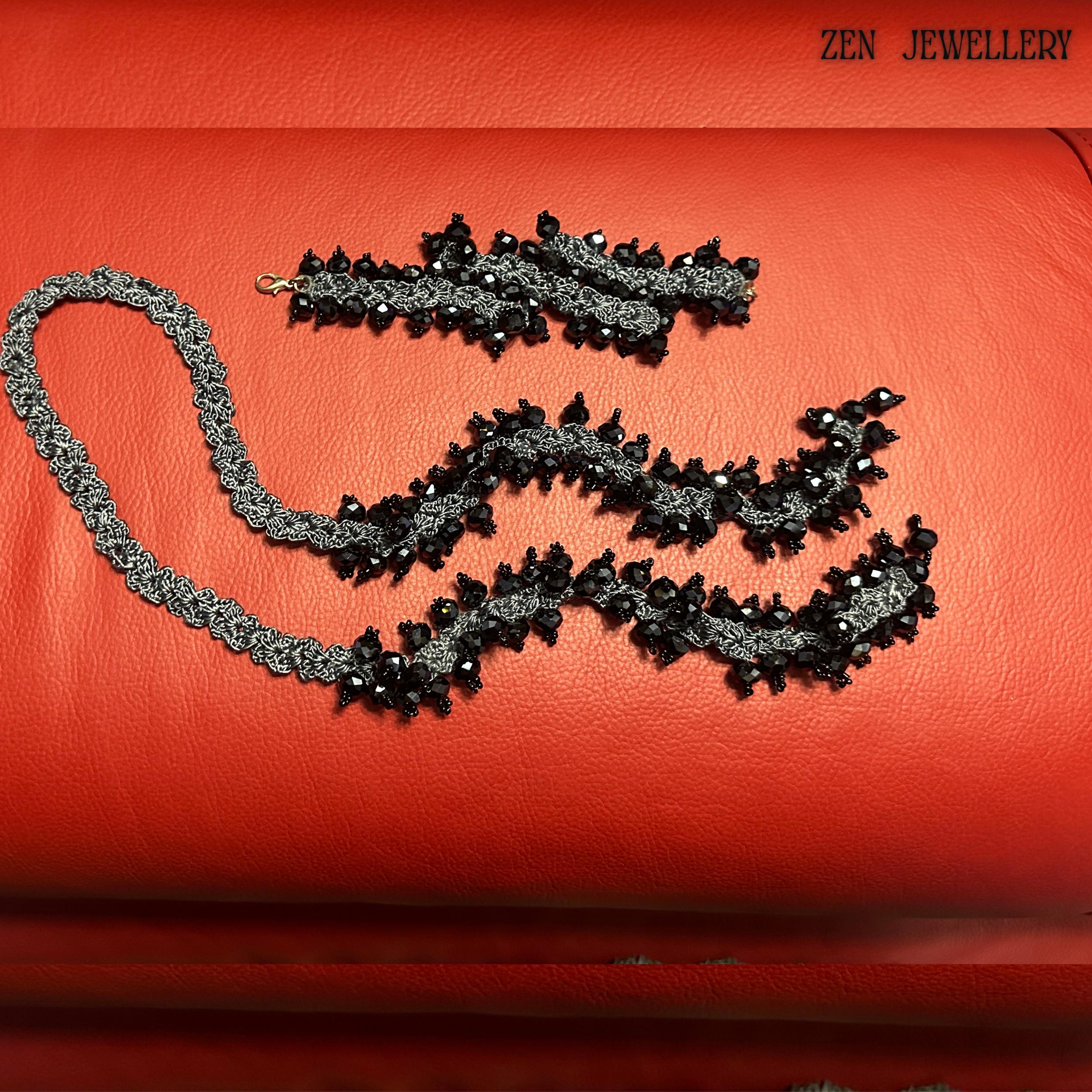 Luxury black crystal jewellery set with oversized faceted beads attached to a charcoal grey crochet silk structure, styled on a rich red background. The necklace and bracelet feature repeating clustered crystal elements, creating a dramatic chunky silhouette while remaining wearable and lightweight. The handcrafted crochet technique adds artistic texture and uniqueness, making every section visually detailed.