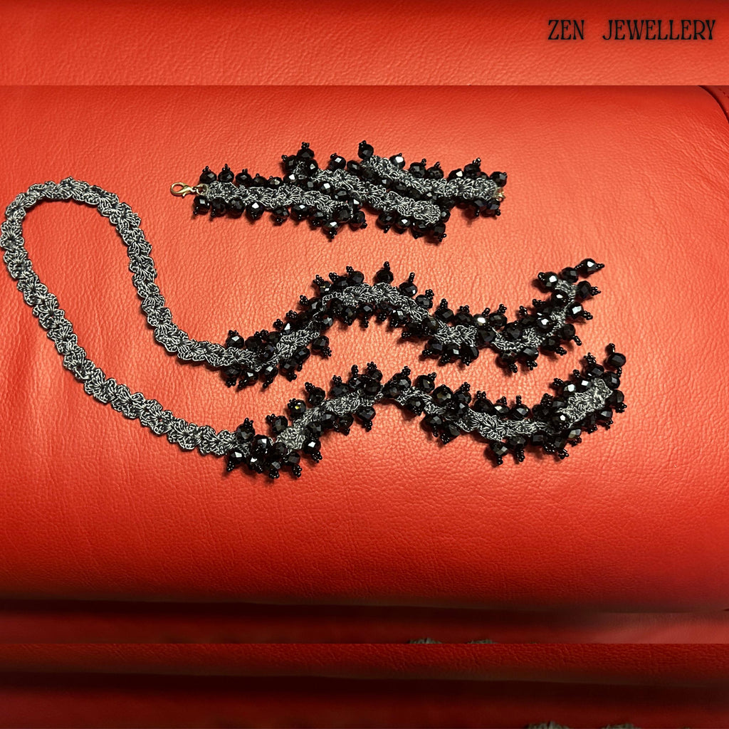 Luxury black crystal jewellery set with oversized faceted beads attached to a charcoal grey crochet silk structure, styled on a rich red background. The necklace and bracelet feature repeating clustered crystal elements, creating a dramatic chunky silhouette while remaining wearable and lightweight. The handcrafted crochet technique adds artistic texture and uniqueness, making every section visually detailed.