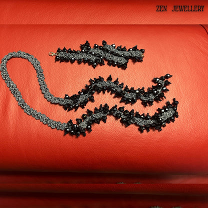 Luxury black crystal jewellery set with oversized faceted beads attached to a charcoal grey crochet silk structure, styled on a rich red background. The necklace and bracelet feature repeating clustered crystal elements, creating a dramatic chunky silhouette while remaining wearable and lightweight. The handcrafted crochet technique adds artistic texture and uniqueness, making every section visually detailed.