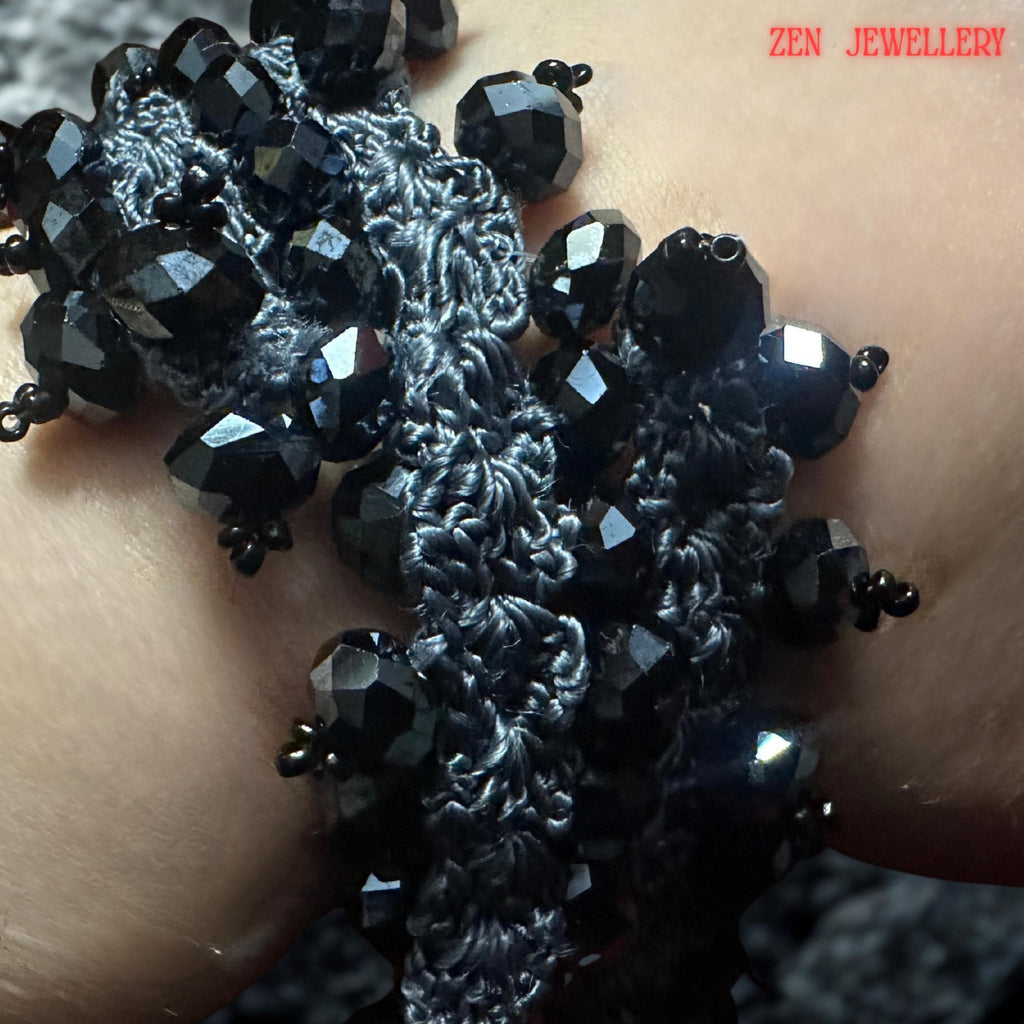 A handcrafted black crystal jewellery set resting on a textured model wrist, featuring charcoal grey crochet silk intertwined with faceted black stones. The necklace and bracelet have an organic, flowing structure with repeating bead clusters creating volume and visual movement. The close-up composition reveals artisan detailing and the lightweight textile-based construction despite the bold chunky visual appearance.