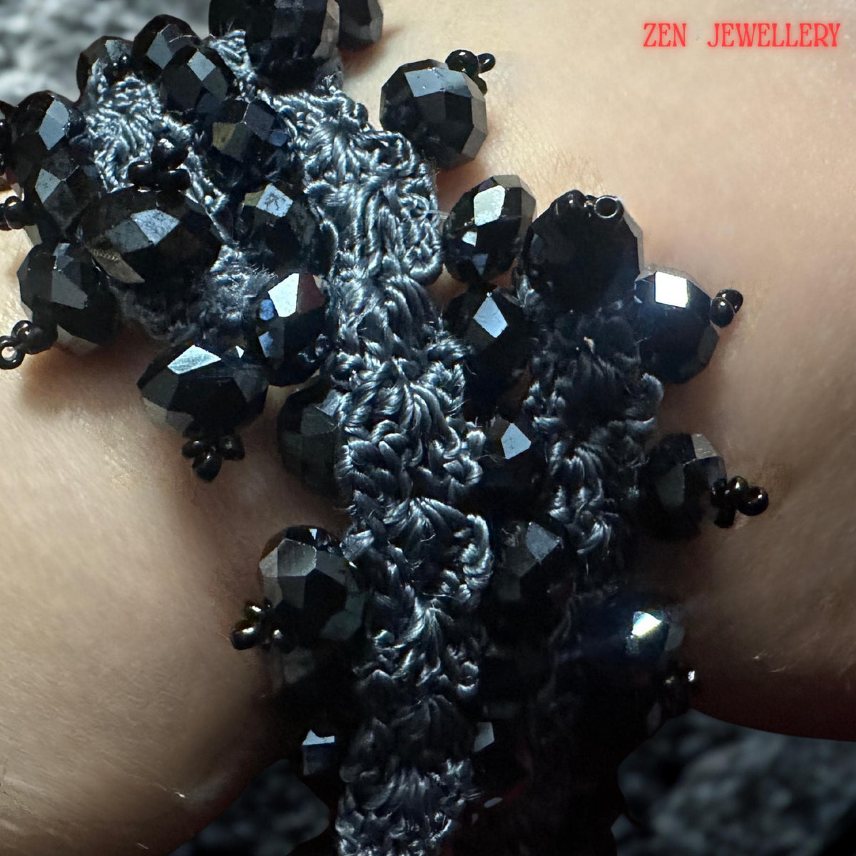 A handcrafted black crystal jewellery set resting on a textured model wrist, featuring charcoal grey crochet silk intertwined with faceted black stones. The necklace and bracelet have an organic, flowing structure with repeating bead clusters creating volume and visual movement. The close-up composition reveals artisan detailing and the lightweight textile-based construction despite the bold chunky visual appearance.