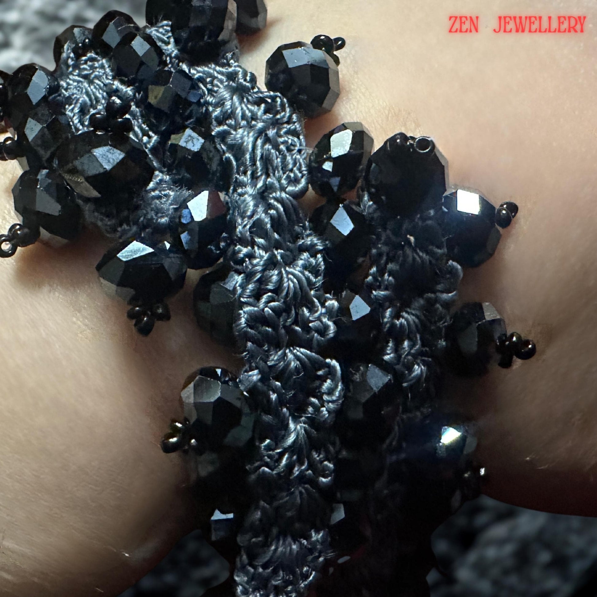 A handcrafted black crystal jewellery set resting on a textured model wrist, featuring charcoal grey crochet silk intertwined with faceted black stones. The necklace and bracelet have an organic, flowing structure with repeating bead clusters creating volume and visual movement. The close-up composition reveals artisan detailing and the lightweight textile-based construction despite the bold chunky visual appearance.