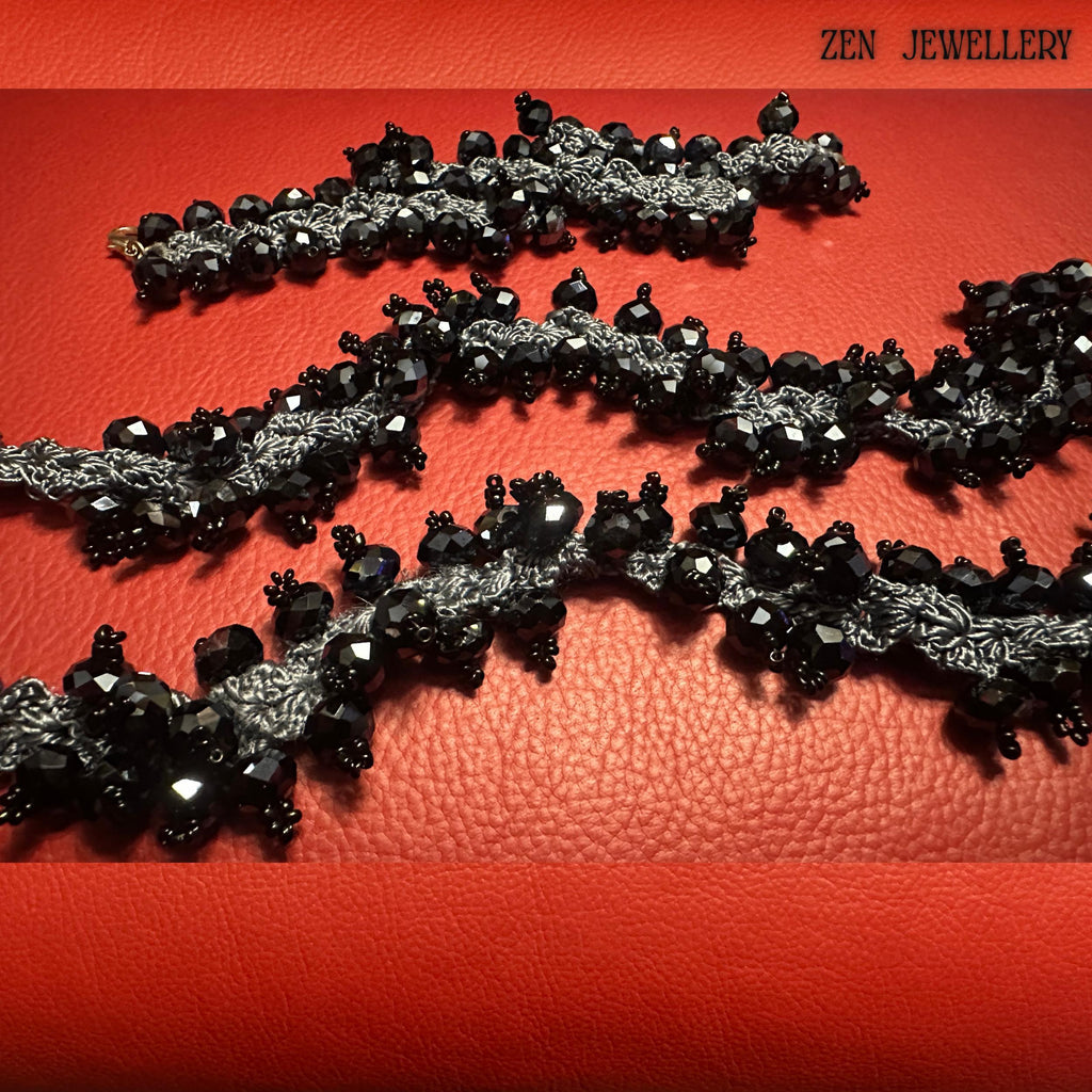 Handmade black crystal necklace displayed partially coiled to demonstrate its flexibility and artisan crochet structure. The faceted black crystals create a reflective glossy surface while the charcoal silk crochet lace introduces softness and handcrafted character. The red background enhances the dramatic contrast of the black jewellery. A visible hand holding part of the necklace helps buyers understand the scale of the oversized chunky stateme