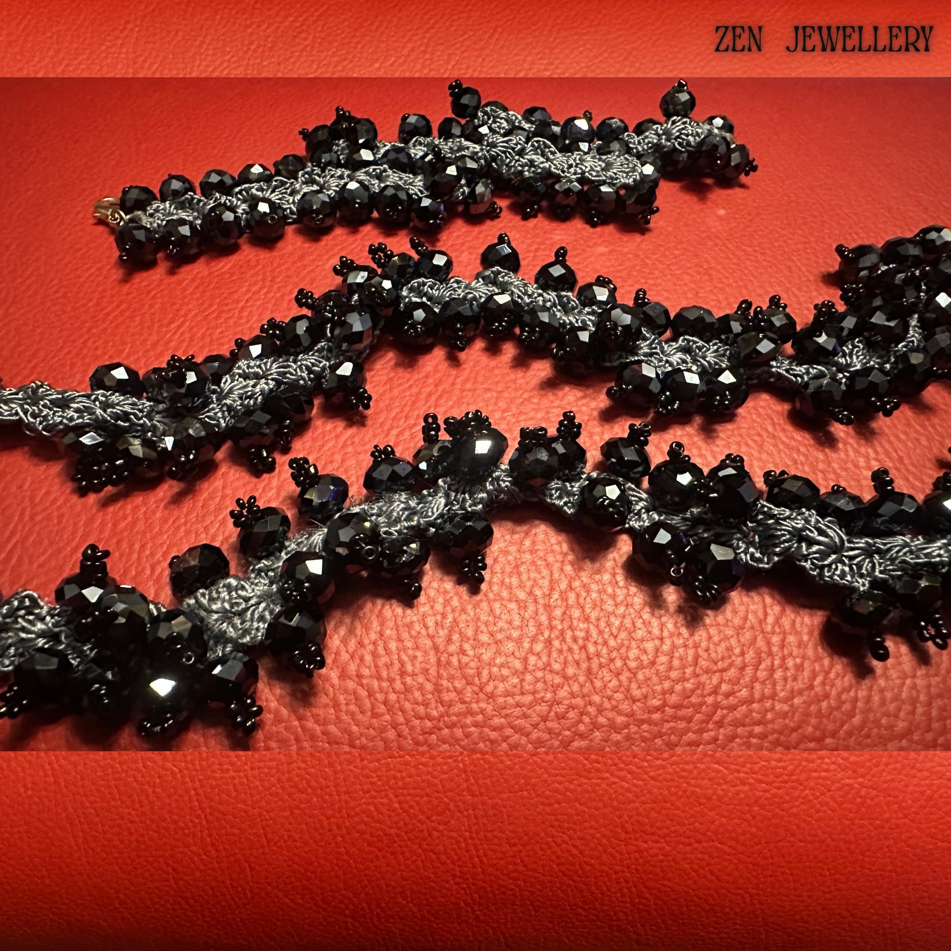 Handmade black crystal necklace displayed partially coiled to demonstrate its flexibility and artisan crochet structure. The faceted black crystals create a reflective glossy surface while the charcoal silk crochet lace introduces softness and handcrafted character. The red background enhances the dramatic contrast of the black jewellery. A visible hand holding part of the necklace helps buyers understand the scale of the oversized chunky stateme