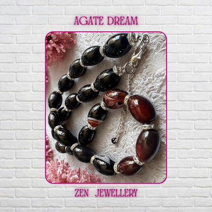 Close-up view of a handmade black agate gemstone necklace arranged on soft textured fabric with delicate pink flowers in the background. Large polished oval beads alternate with striped burgundy agate stones and shimmering rhinestone spacer rings. The silver clasp and adjustable chain add elegant finishing details. The necklace has a smooth glossy surface and luxurious reflective shine. Necklace measures 47 cm or approximately 18.5 inches long.