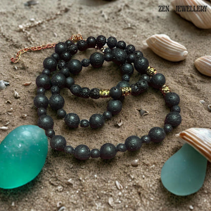 Matching lava stone bracelet shown with matte black volcanic beads and decorative gold spacer accents. The porous natural lava stones create an earthy bohemian aesthetic while the polished golden details add elegance and sophistication. The set displayed on sand next to green stones.