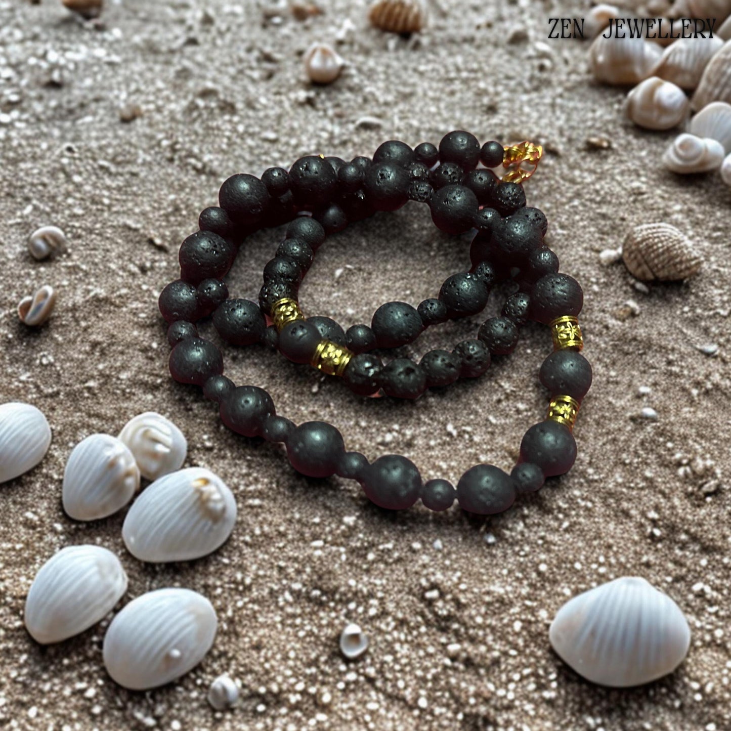 Elegant close-up of the necklace clasp and golden extension chain highlighting the refined handmade details of the volcanic bead jewellery set. The natural lava stones feature a porous matte surface with organic texture variations that make each piece unique. The combination of black volcanic beads and warm gold accents creates a balanced minimalist bohemian style. Jewellery displayed on snad next to shells