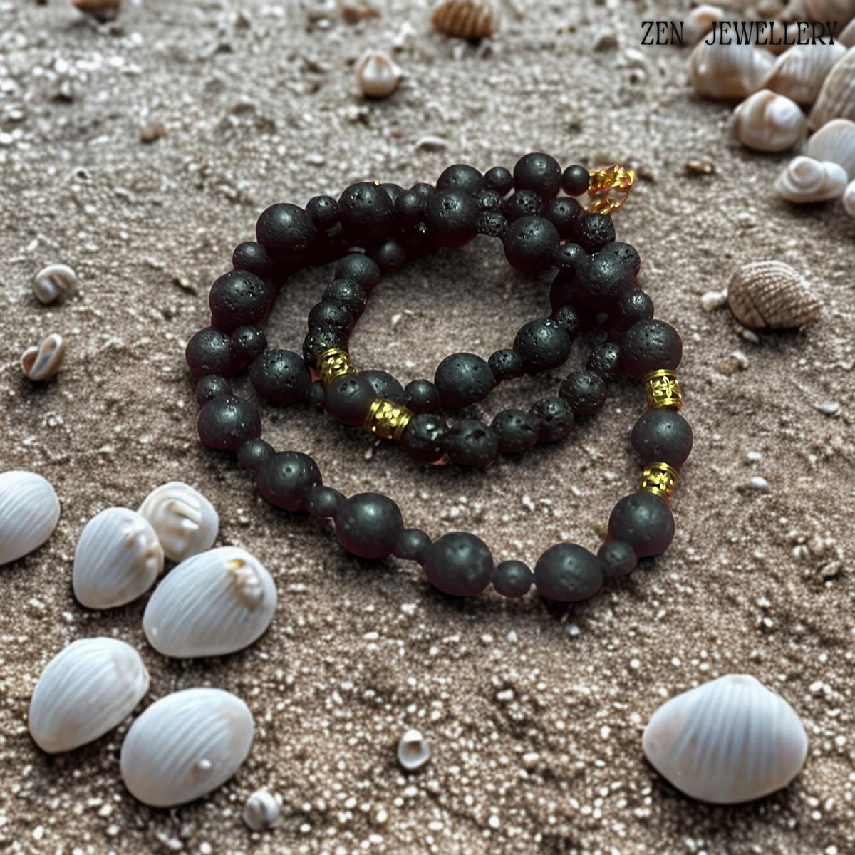 Elegant close-up of the necklace clasp and golden extension chain highlighting the refined handmade details of the volcanic bead jewellery set. The natural lava stones feature a porous matte surface with organic texture variations that make each piece unique. The combination of black volcanic beads and warm gold accents creates a balanced minimalist bohemian style. Jewellery displayed on snad next to shells