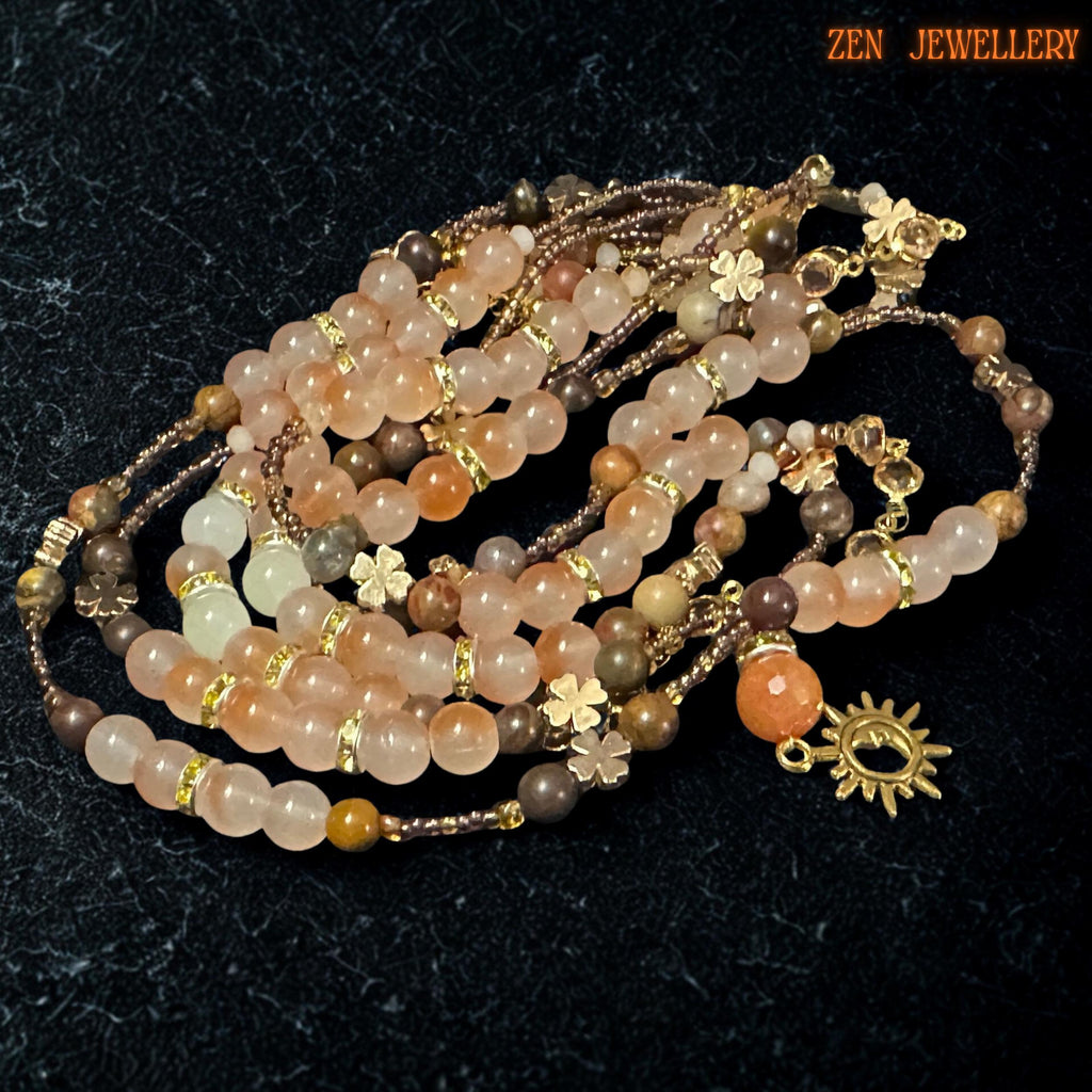 Celestial bohemian jewellery set featuring a handcrafted layered necklace and bracelet with a warm, earthy gemstone palette including blush orange aventurine, beige jade-style beads, champagne crystal accents, and gold chain details. Dark stone background highlights the sparkle of faceted beads and luxurious golden tones while floral charms add delicate, feminine texture.