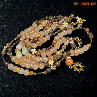 Celestial bohemian jewellery set featuring a handcrafted layered necklace and bracelet with a warm, earthy gemstone palette including blush orange aventurine, beige jade-style beads, champagne crystal accents, and gold chain details. Dark stone background highlights the sparkle of faceted beads and luxurious golden tones while floral charms add delicate, feminine texture.
