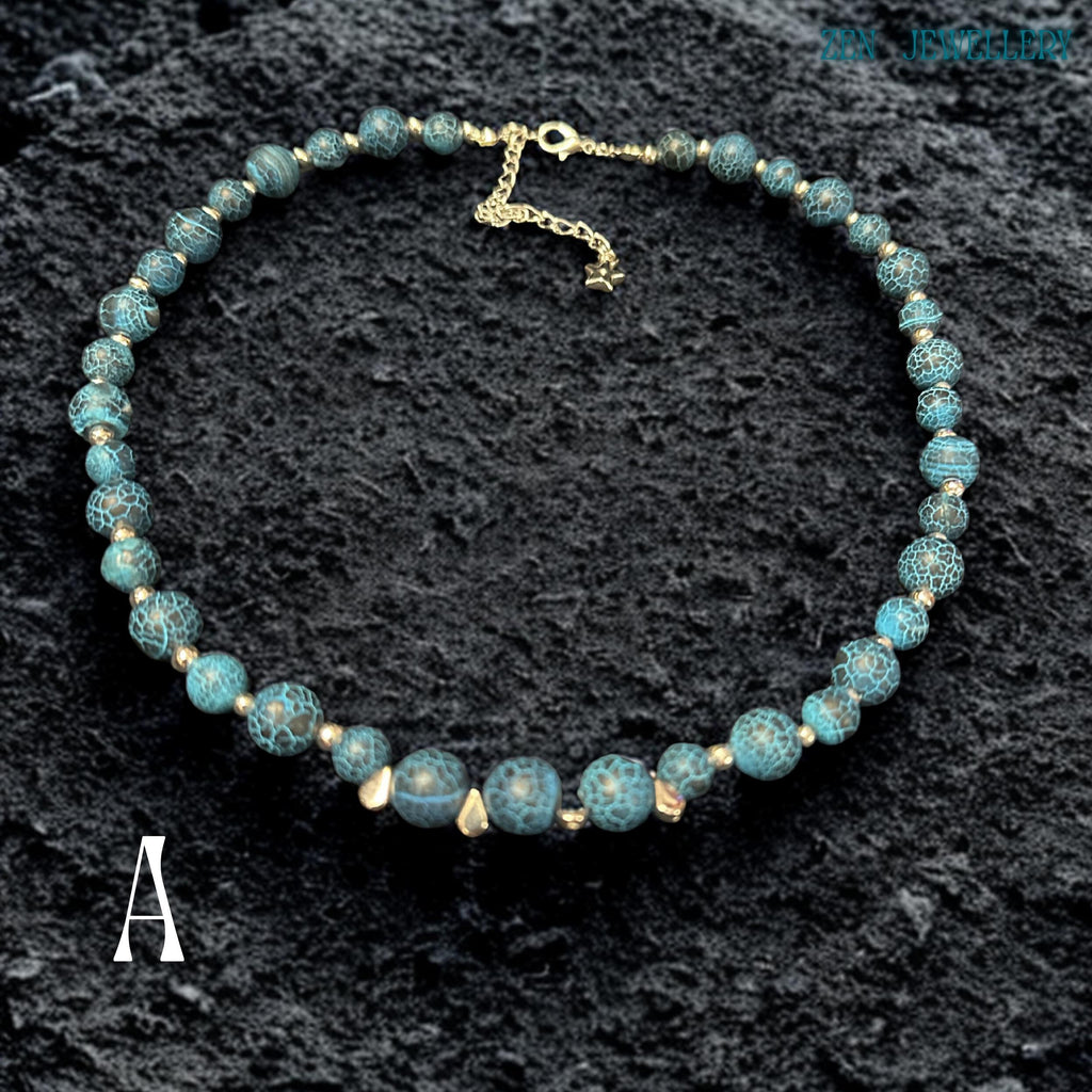 Close-up image of Blue Crackle Agate gemstone beads showing detailed crackled vein textures in deep turquoise, black, and ocean blue shades separated by polished silver spacer beads that reflect light softly against the dark background, highlighting the natural stone patterns, handcrafted jewellery construction, layered bead arrangement, and elegant celestial styling details that create a luxurious bohemian appearance.