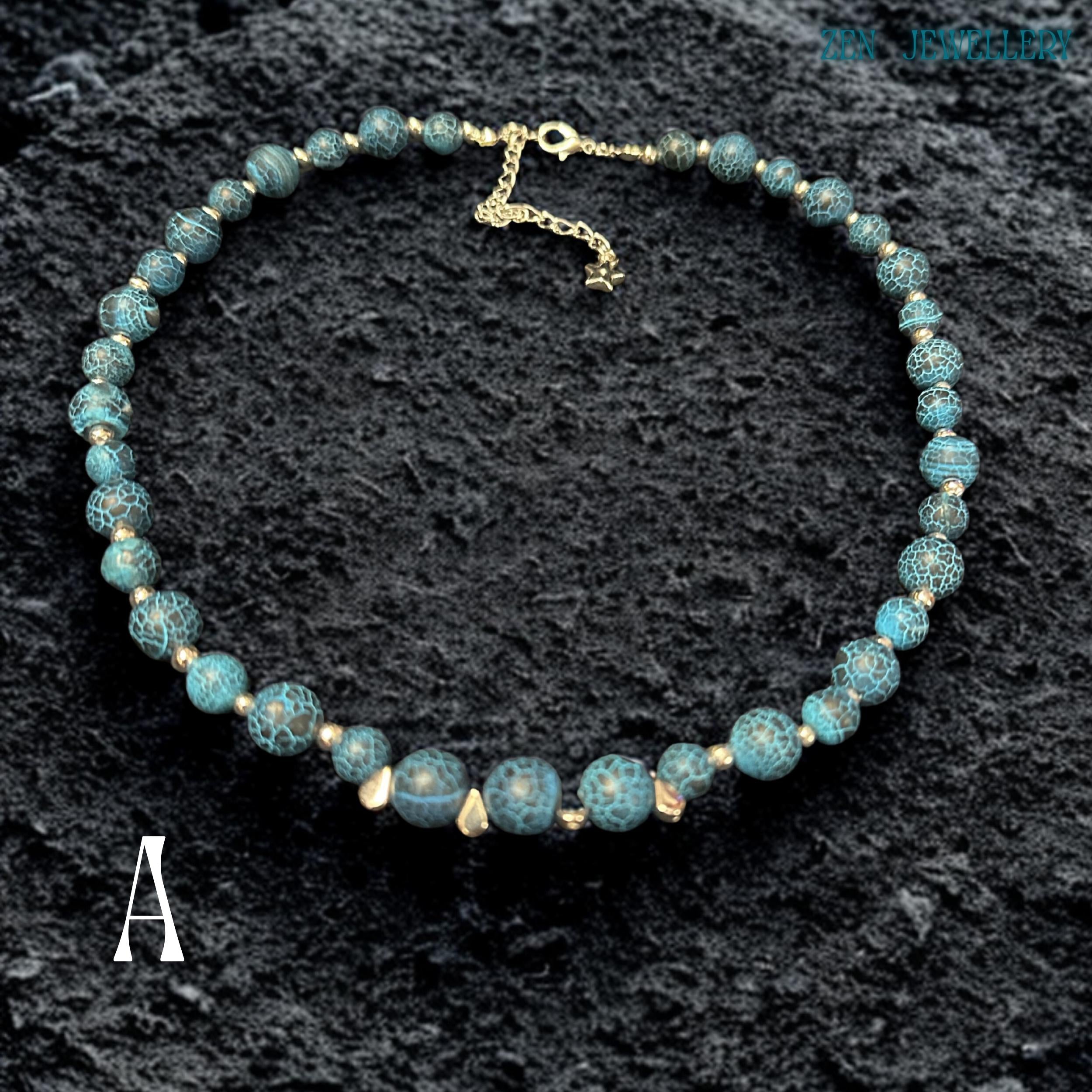 Close-up image of Blue Crackle Agate gemstone beads showing detailed crackled vein textures in deep turquoise, black, and ocean blue shades separated by polished silver spacer beads that reflect light softly against the dark background, highlighting the natural stone patterns, handcrafted jewellery construction, layered bead arrangement, and elegant celestial styling details that create a luxurious bohemian appearance.
