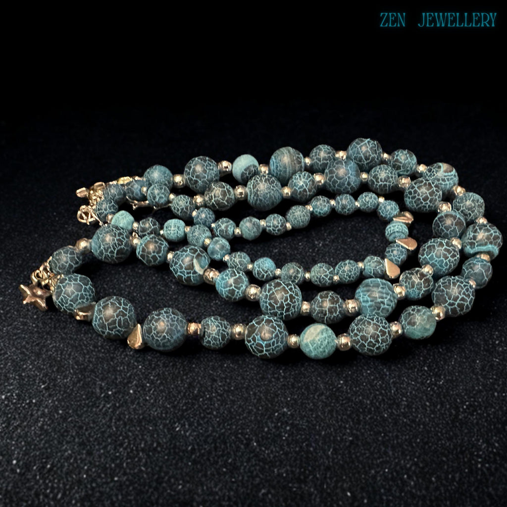 Macro close-up photograph of the Blue Crackle Agate gemstone surface revealing intricate natural crackled vein patterns across polished turquoise and black beads separated by reflective silver spacers that create a luxurious gemstone texture full of earthy character, artistic depth, and handcrafted bohemian elegance suitable for statement jewellery lovers and gemstone collectors.