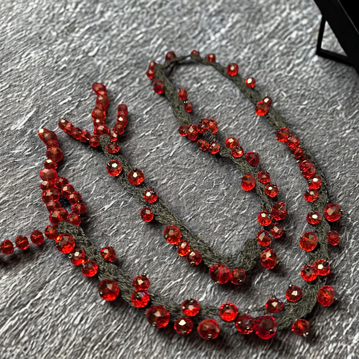 Close-up image showing detailed faceted RED CRYSTAL beads attached carefully onto black crochet strands with handcrafted precision. The rich crimson crystals vary in size and reflect warm light from multiple angles, adding glamorous sparkle and dimensional texture. The soft braided base curves naturally and enhances the organic artistic shape of the necklace. The dramatic colour contrast between red and black gives the jewellery set a luxurious g