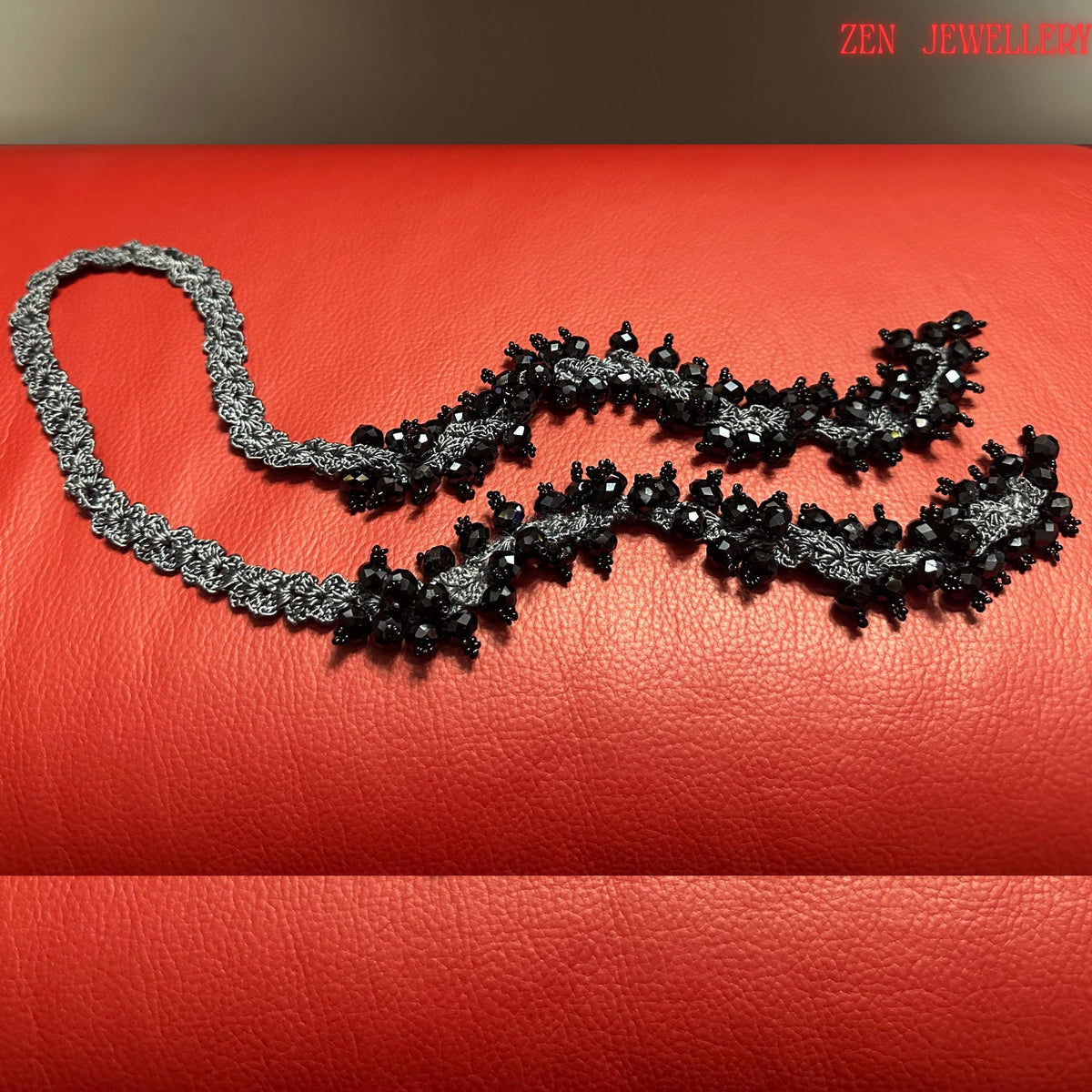 Handcrafted black crystal statement necklace featuring glossy faceted black beads woven into charcoal silk crochet patterns with sterling silver fastening details. The jewellery curves naturally across the surface, showing flexibility and movement. The handmade crochet lace texture contrasts beautifully with the polished crystal surfaces, giving the piece an elegant evening jewellery aesthetic suitable for black dresses and formal outfits.