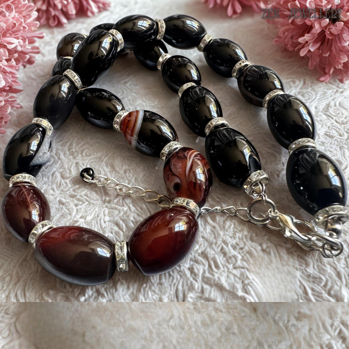 Close detailed photograph of AGATE GEMSTONE jewellery necklace with smooth oval black beads and dark burgundy agate stones featuring natural striped patterns. Silver rhinestone spacers catch the light beautifully and create glamorous sparkle between the gemstones. The necklace includes an adjustable silver chain and secure clasp. Total necklace length is 47 cm or around 18.5 inches.