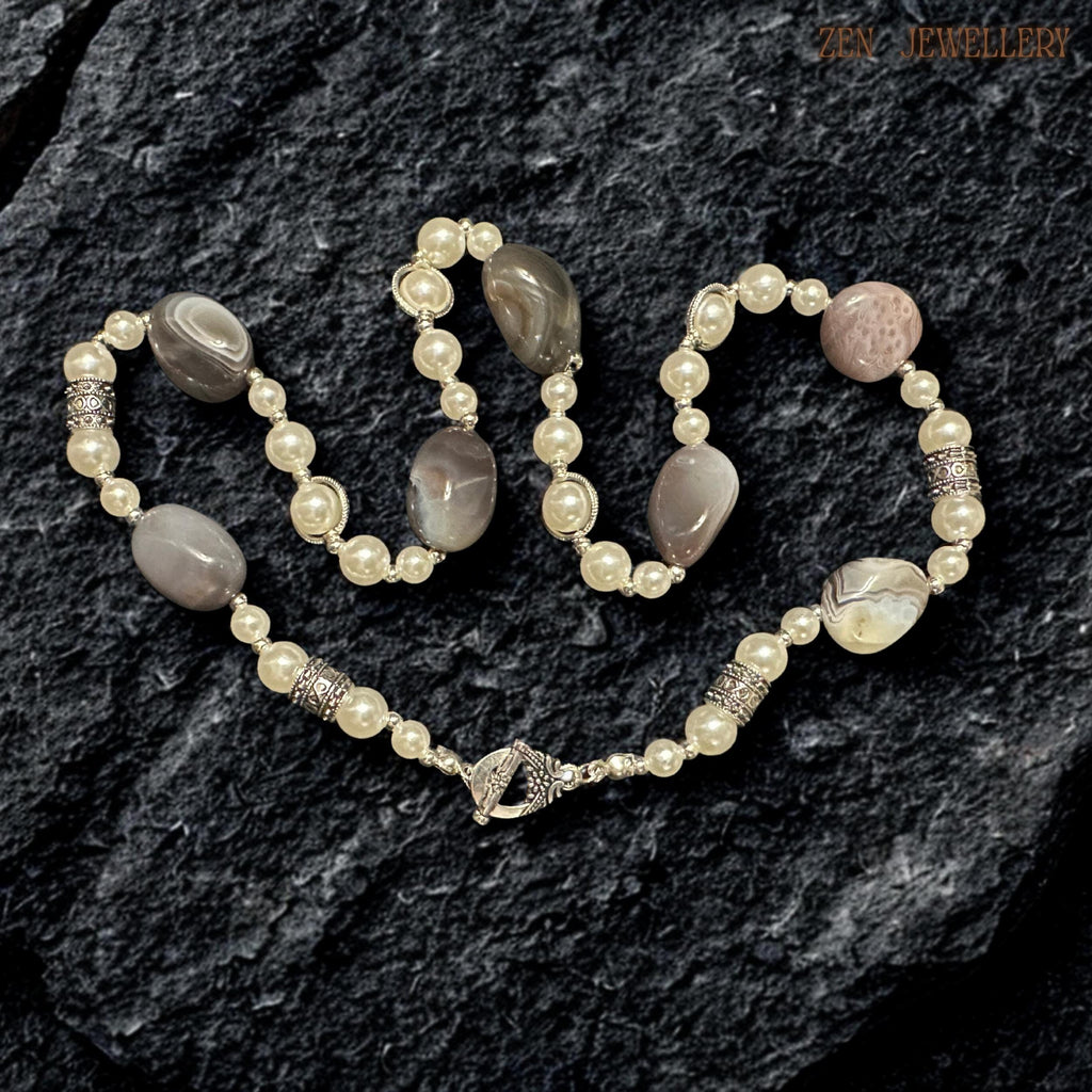 Artistic BOTSWANA AGATE statement necklace featuring natural oval agate gemstones, ivory pearl beads, and detailed silver accents arranged in an elegant layered composition. The striped agate patterns create organic texture and earthy character while the pearls bring classic softness and femininity. Decorative silver rhinestone spacers reflect light delicately and enhance the luxurious handcrafted appearance.