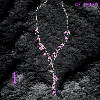 Handmade silver chain necklace with dangling pink and purple gemstone bead charms displayed against a soft blush pink dress and blonde hair. The layered design includes chunky silver chain sections combined with delicate bead strands arranged asymmetrically for a flowing artistic appearance. Polished pastel gemstone beads reflect light softly while faceted crystal accents add sparkle and elegance.