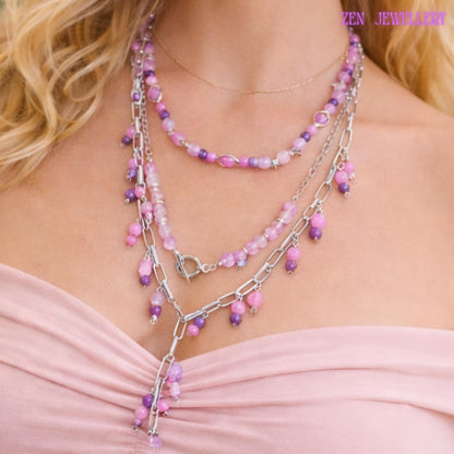 Layered pastel gemstone necklace featuring three silver chain strands decorated with pink, lilac, lavender, and purple bead clusters attached along chunky metallic links. The necklace combines polished round gemstone beads with faceted crystal accents for subtle sparkle and luxurious detail. Small dangling bead charms create movement while the silver toggle clasp becomes part of the decorative front design for a modern handcrafted finish.