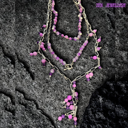 Romantic bohemian necklace displayed on a blonde model wearing a pale pink off shoulder dress, highlighting the layered silver chain design decorated with soft pastel gemstone beads. The necklace features dangling pink and purple bead charms attached along chunky silver chain links, creating graceful movement and playful texture. The combination of polished glossy beads and bold metallic chains creates a modern feminine statement jewellery style.