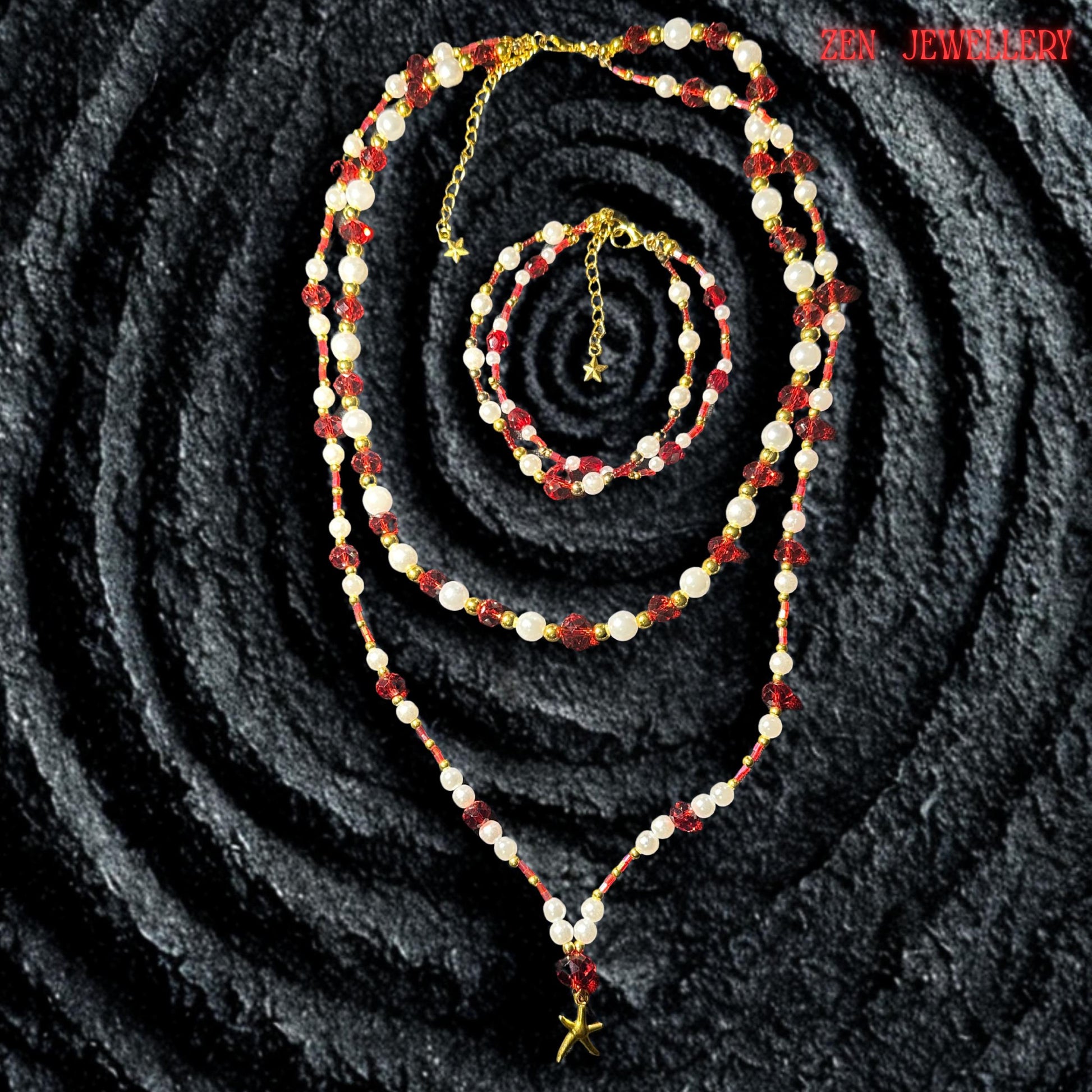 The long layered necklace is arranged in a soft V shape on a dark volcanic textured background highlighting the contrast between the sparkling red crystals and creamy white beads. The lower strand features a delicate golden starfish charm suspended from a ruby red crystal focal bead creating a coastal bohemian style inspired by summer evenings beach fashion and romantic handmade jewellery collections.
