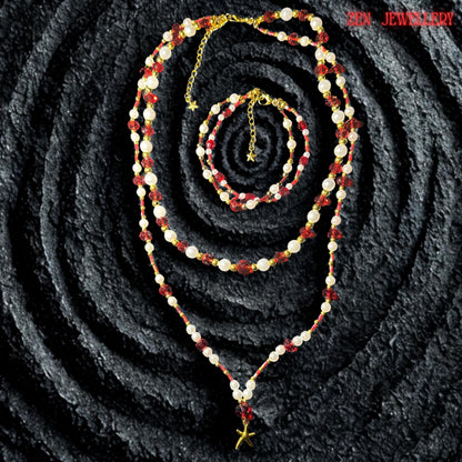 The long layered necklace is arranged in a soft V shape on a dark volcanic textured background highlighting the contrast between the sparkling red crystals and creamy white beads. The lower strand features a delicate golden starfish charm suspended from a ruby red crystal focal bead creating a coastal bohemian style inspired by summer evenings beach fashion and romantic handmade jewellery collections.