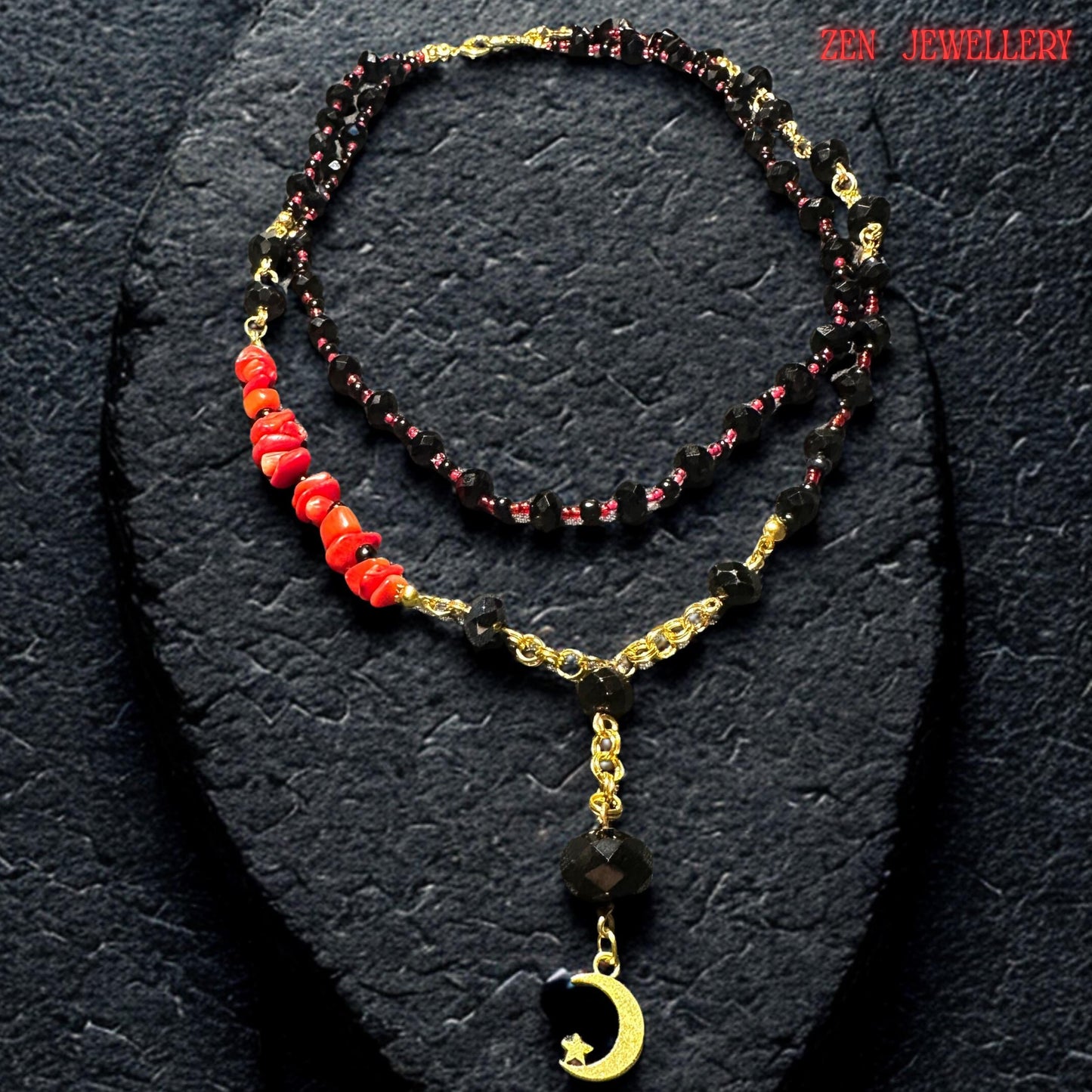 Elegant black onyx style necklace photographed flat on textured dark background showing the layered arrangement of faceted black crystal beads, tiny red accent beads, and polished gold connectors. The moon pendant detail adds a celestial touch while the red coral stones create a striking contrast against the glossy black crystals. Necklace lengths are approximately 17.3 inches and 20.4 inches.