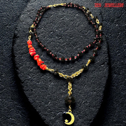 Elegant black onyx style necklace photographed flat on textured dark background showing the layered arrangement of faceted black crystal beads, tiny red accent beads, and polished gold connectors. The moon pendant detail adds a celestial touch while the red coral stones create a striking contrast against the glossy black crystals. Necklace lengths are approximately 17.3 inches and 20.4 inches.