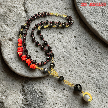 Fashion jewellery flat lay showing a layered black crystal necklace with glossy faceted beads, tiny deep red accent crystals, and natural red coral stones arranged on a textured dark background. The gold moon pendant hanging beneath the central bead adds symbolic meaning associated with intuition and feminine energy. The layered necklace style creates elegant movement and dimension.