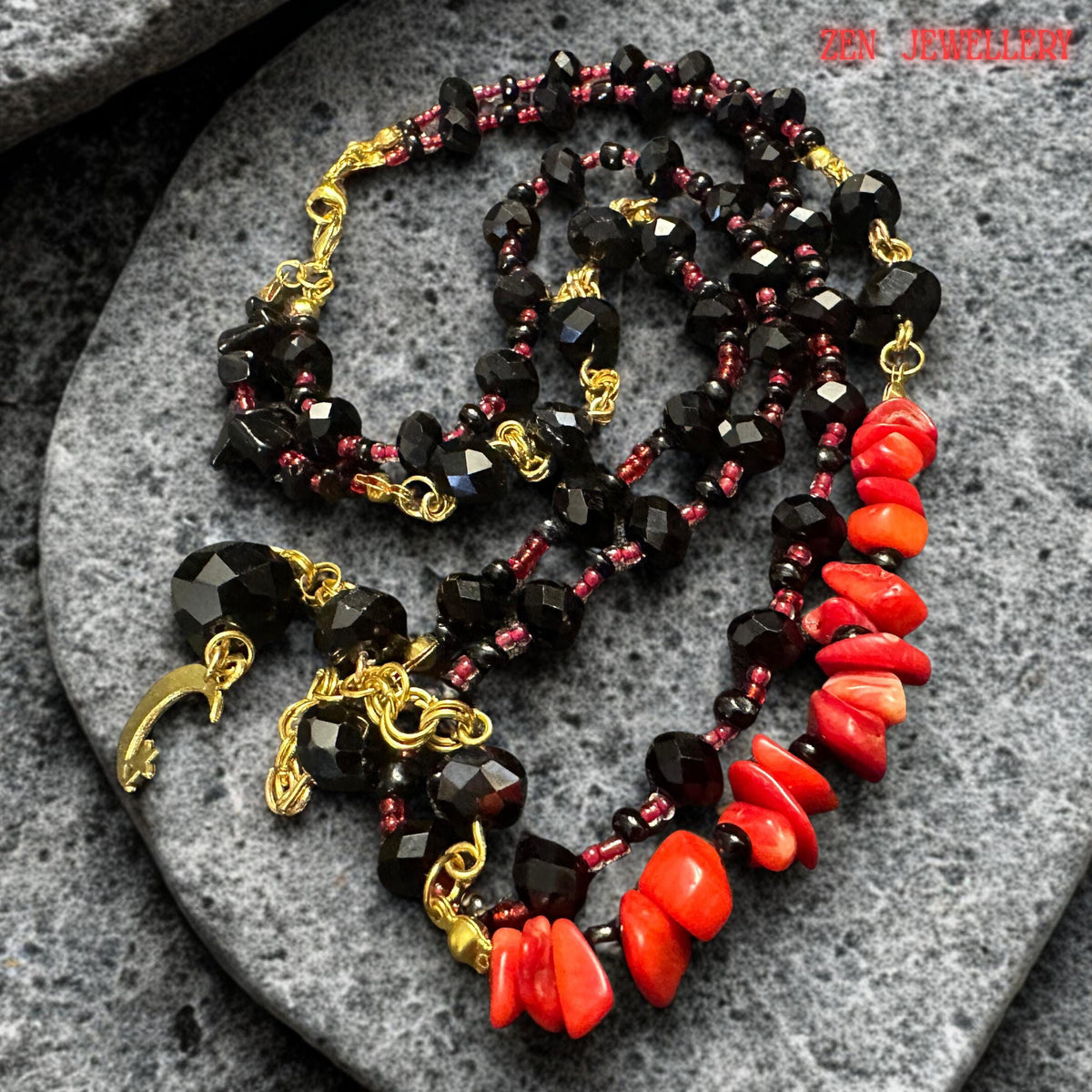 Full necklace display showing two layered strands of glossy black beads accented with gold details and vibrant red coral stones. The asymmetrical arrangement creates a unique handcrafted appearance while the moon pendant centrepiece gives the necklace mystical symbolism and dramatic visual appeal. The necklace is suitable for evening styling or statement fashion looks.