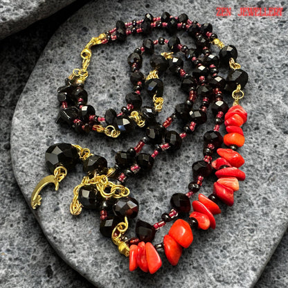 Full necklace display showing two layered strands of glossy black beads accented with gold details and vibrant red coral stones. The asymmetrical arrangement creates a unique handcrafted appearance while the moon pendant centrepiece gives the necklace mystical symbolism and dramatic visual appeal. The necklace is suitable for evening styling or statement fashion looks.
