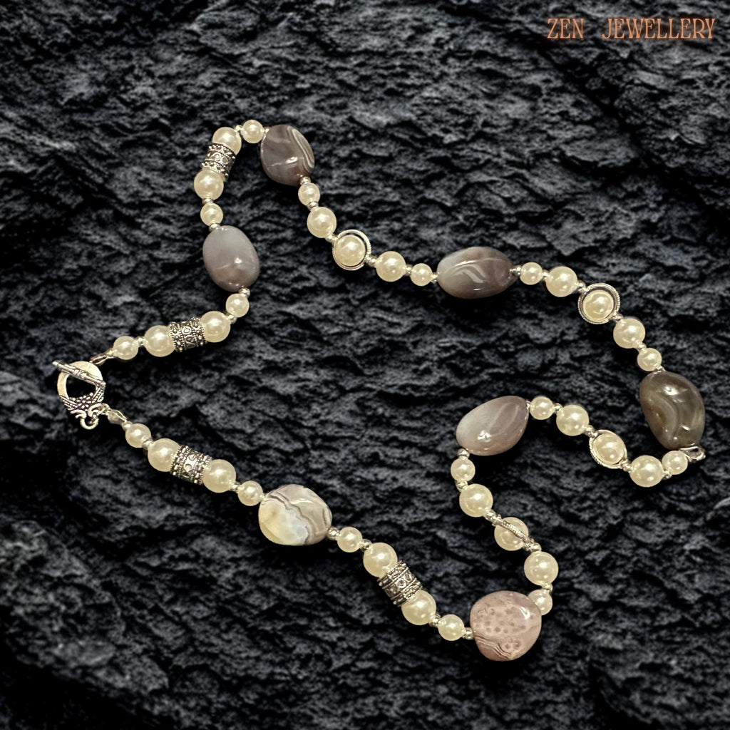 Elegant gemstone jewellery photographed on a dark textured surface to emphasise the soft neutral beauty of the BOTSWANA AGATE stones and creamy pearl details. The necklace combines polished agate focal beads with smooth pearls and silver toggle closure elements for a timeless artistic appearance. The layered arrangement creates graceful balance while silver rhinestone accents add delicate sparkle and vintage charm.